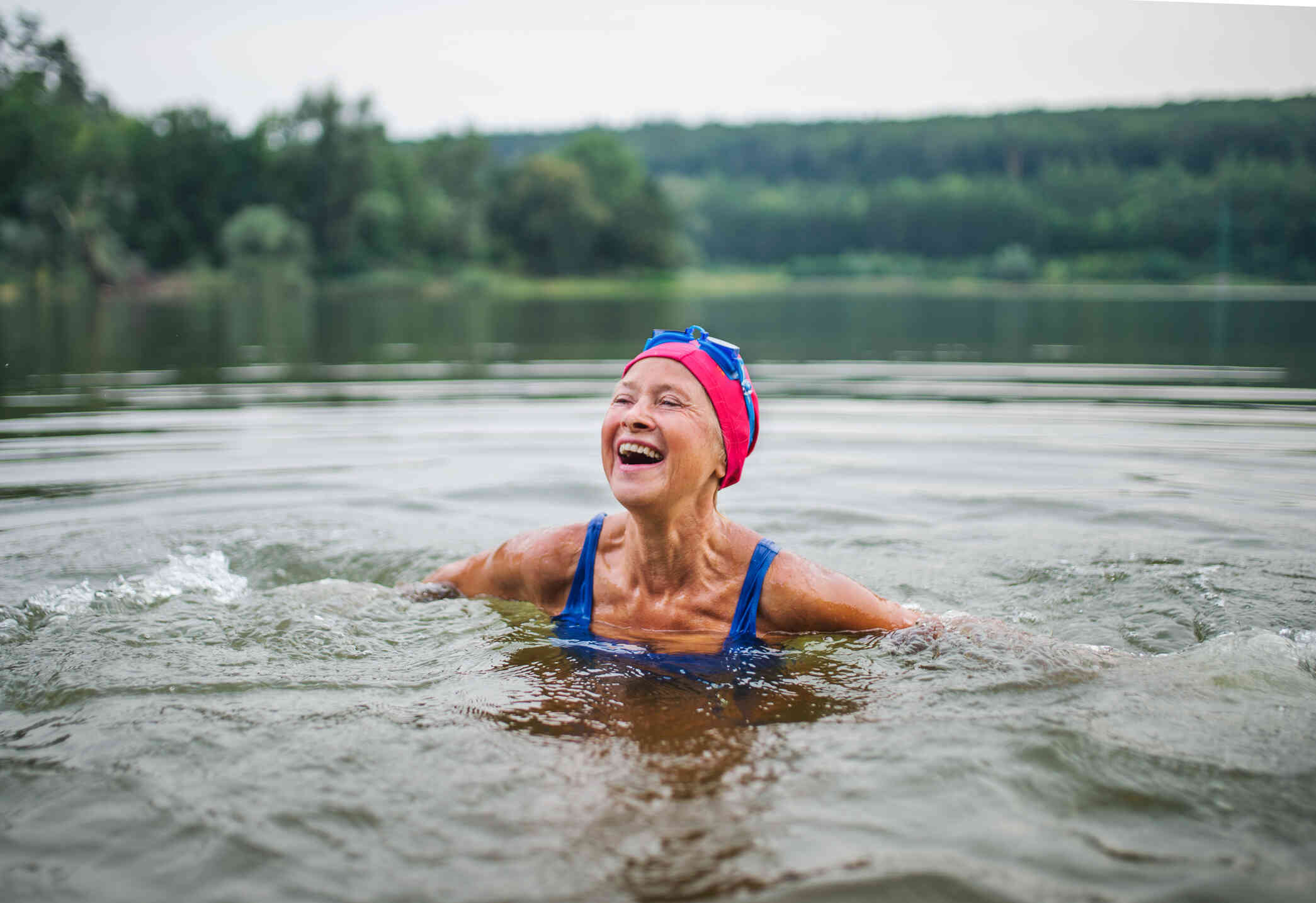 An elderly woman in a pink swimcap smiles brightly while swimming in a lake on a sunny day.